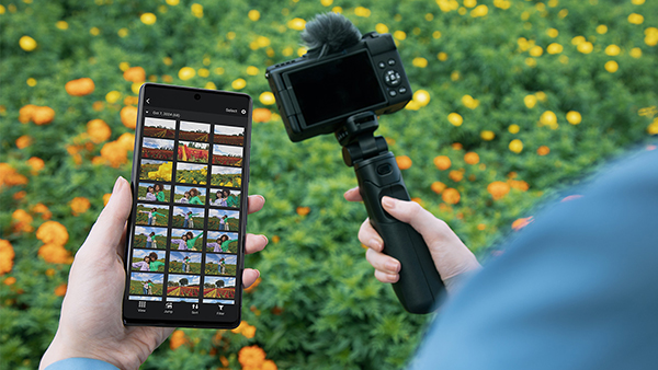 This is an image of the Canon PowerShot V1 and a smartphone, which are both held by someone wearing a blue shirt with orange flowers in the background.