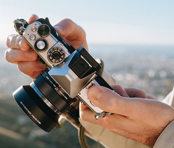 A photographer's hands are holding the Olympus O-M3 mirrorless camera with the Olympus M.ZUIKO DIGITAL 17mm F1.8 II lens attached to the camera body.