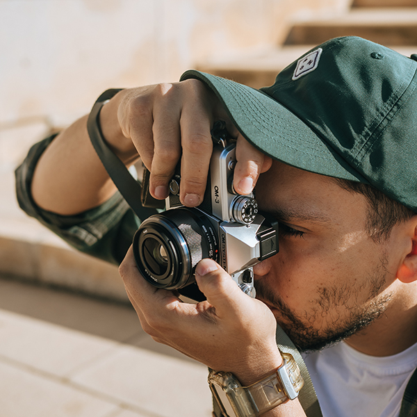 A photographer is holding the Olympus O-M3 mirrorless camera with the Olympus M.ZUIKO DIGITAL 25mm F1.8 II lens attached to the camera body.