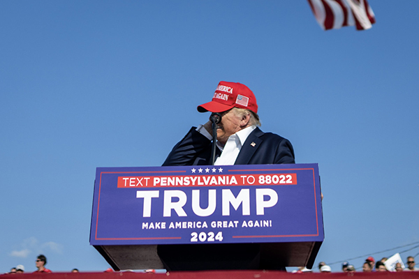 This is a photograph by Doug Mills, which shows President Donald Trump speaking at a microphone and a bullet, which is depicted as a line, to his left.