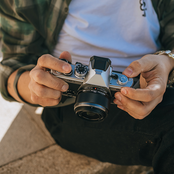 A photographerâ€™s hands are holding the Olympus O-M3 mirrorless camera with the Olympus M.ZUIKO DIGITAL 25mm F1.8 II lens attached to the camera body.