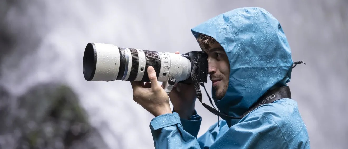  A photographer in a blue hooded rain jacket holding a camera with an OM SYSTEM M.Zuiko Digital ED 50-200mm f/2.8 IS PRO. 